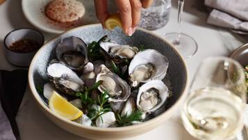 A bowl of oysters garnished with lemon and herbs, a glass of white wine, and a small bowl of seasoning on a dining table.