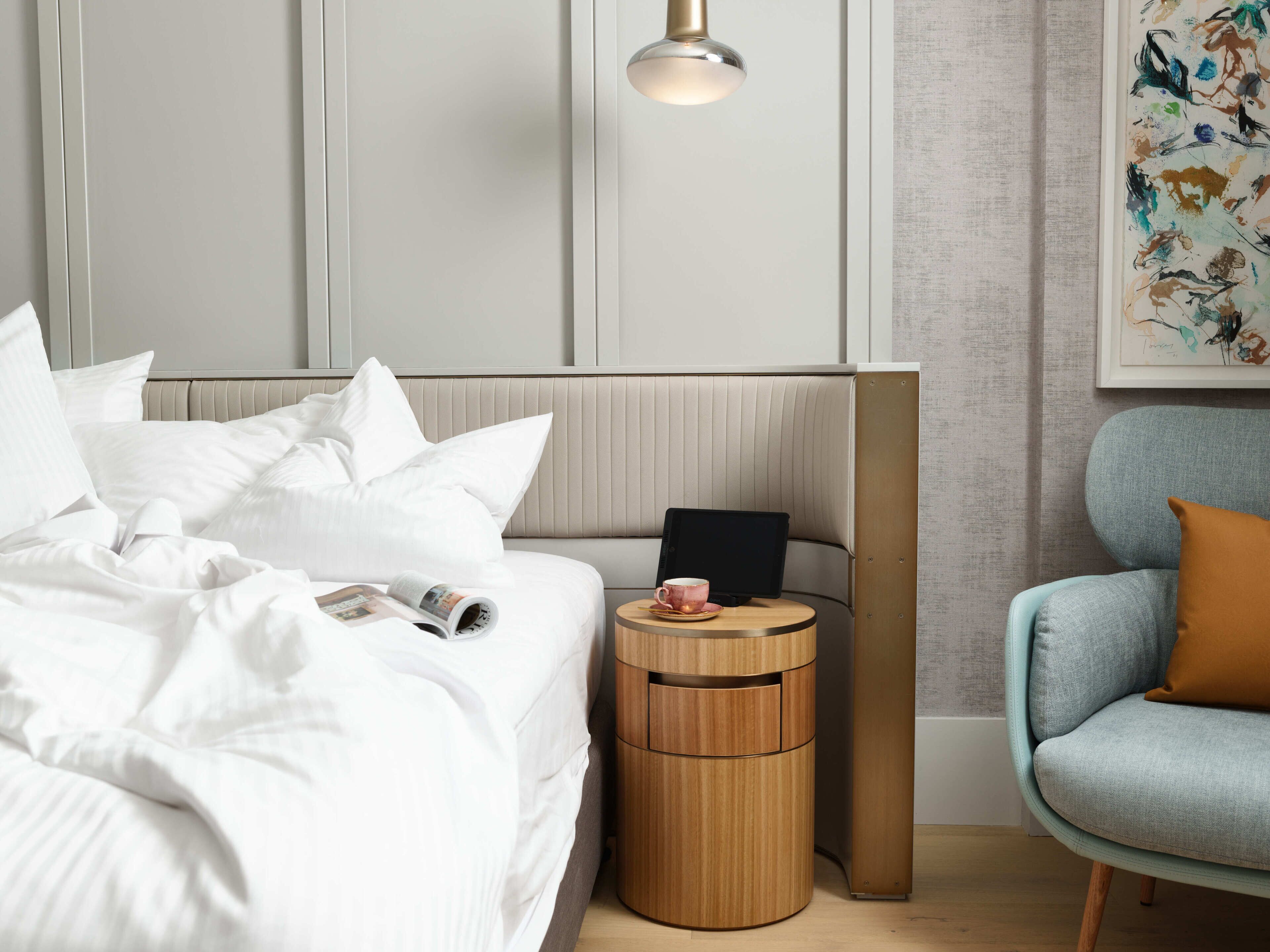 A neatly made bed with white linens, a bedside table with a tablet and a cup, a blue armchair with a brown cushion, and a wall-mounted lamp.