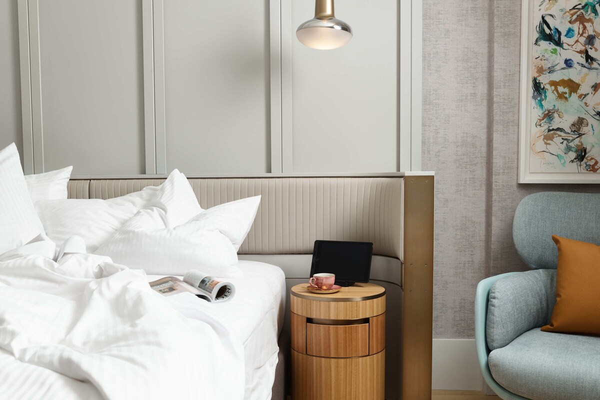 Un lit bien fait, avec du linge de lit blanc, une table de chevet sur laquelle se trouvent une tablette et une tasse, un fauteuil bleu avec un coussin marron et une lampe fixée au mur.