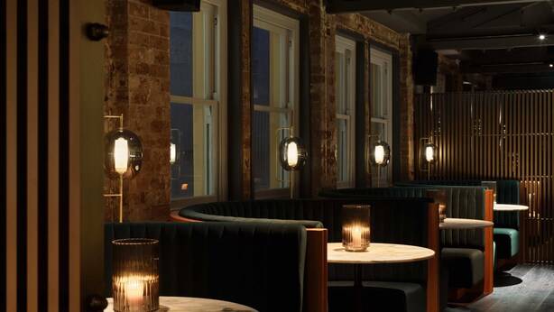 Un restaurant à la lumière tamisée, avec des murs en briques, de grandes fenêtres et des tables rondes entourées de tabourets.