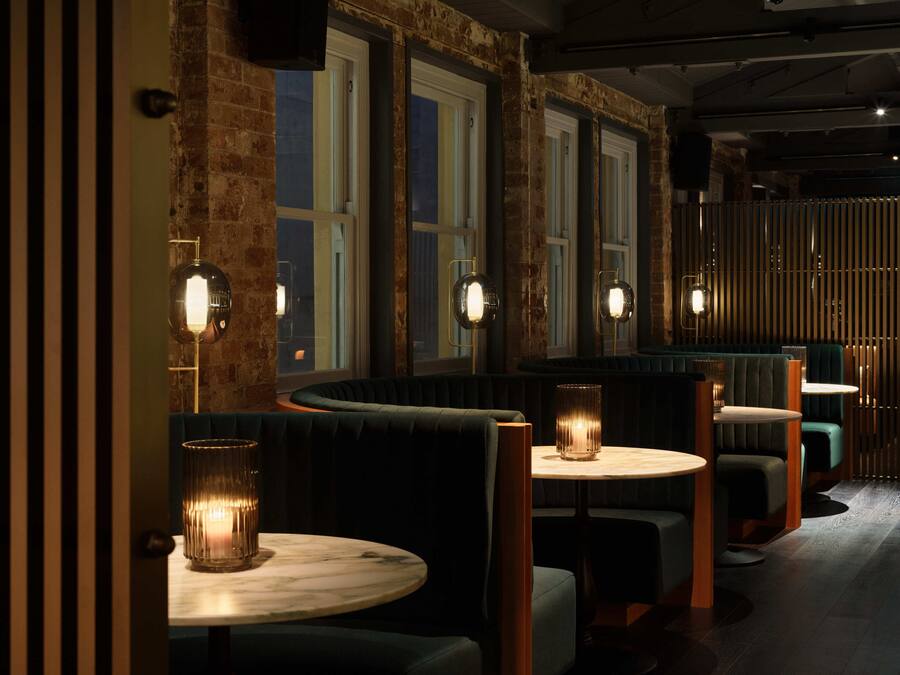 Un restaurante tenuemente iluminado con paredes de ladrillo, ventanas amplias y mesas redondas con taburetes.
