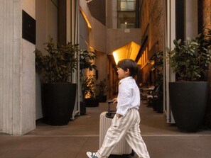 A child walking with a suitcase in a corridor with large potted plants and a warm interior lighting.