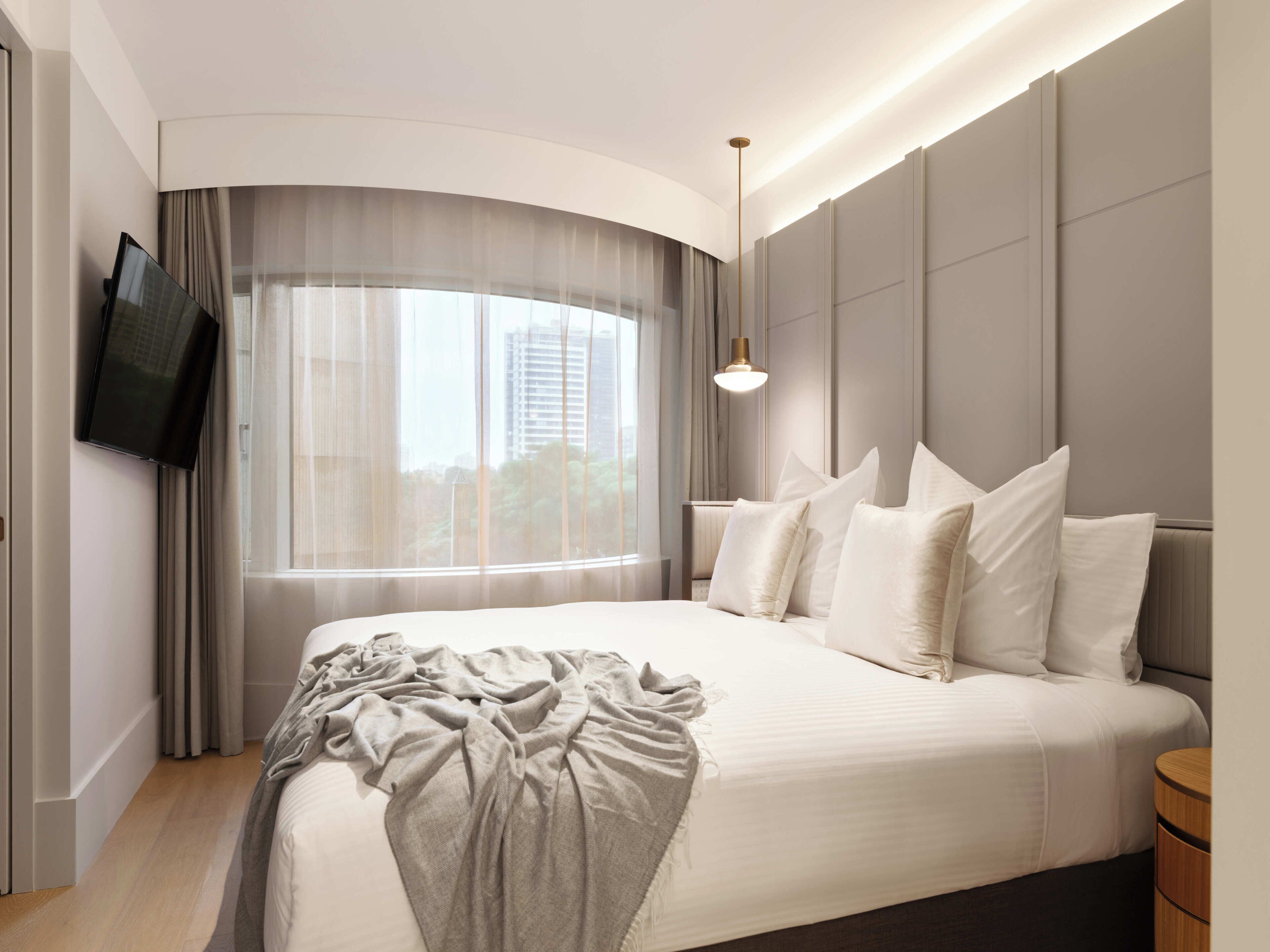 A modern hotel room with a large bed, a flat-screen TV, a bedside lamp, and a view of the city through sheer curtains.