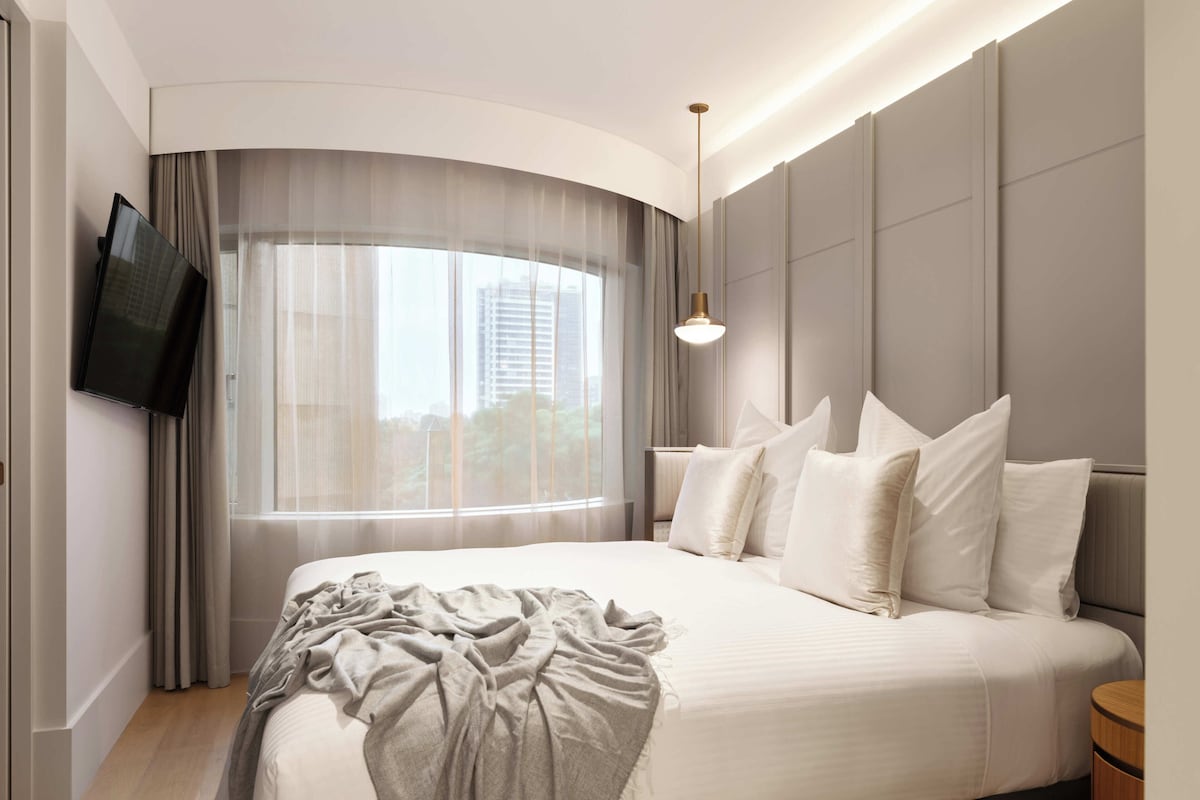 Une chambre d’hôtel moderne dotée d’un grand lit, d’un téléviseur à écran plat, d’une lampe de chevet et offrant une vue sur la ville à travers de fines rideaux.
