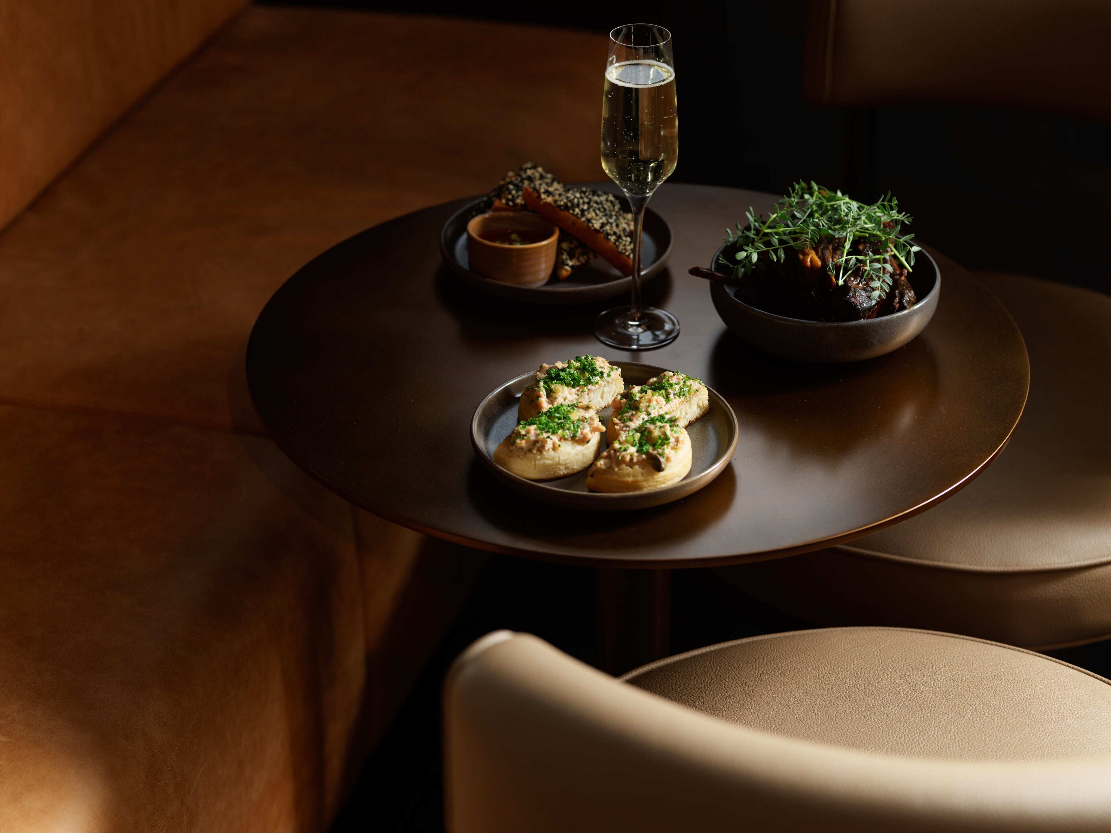 A round table with a glass of champagne, a bowl of greens, and a plate of appetizers.