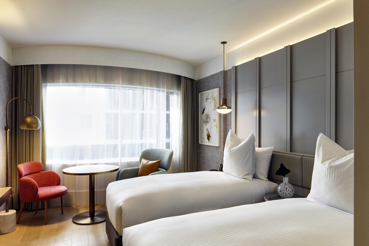 Een moderne hotelkamer met twee bedden, een ronde tafel, een rode stoel en een groot raam met dunne gordijnen.