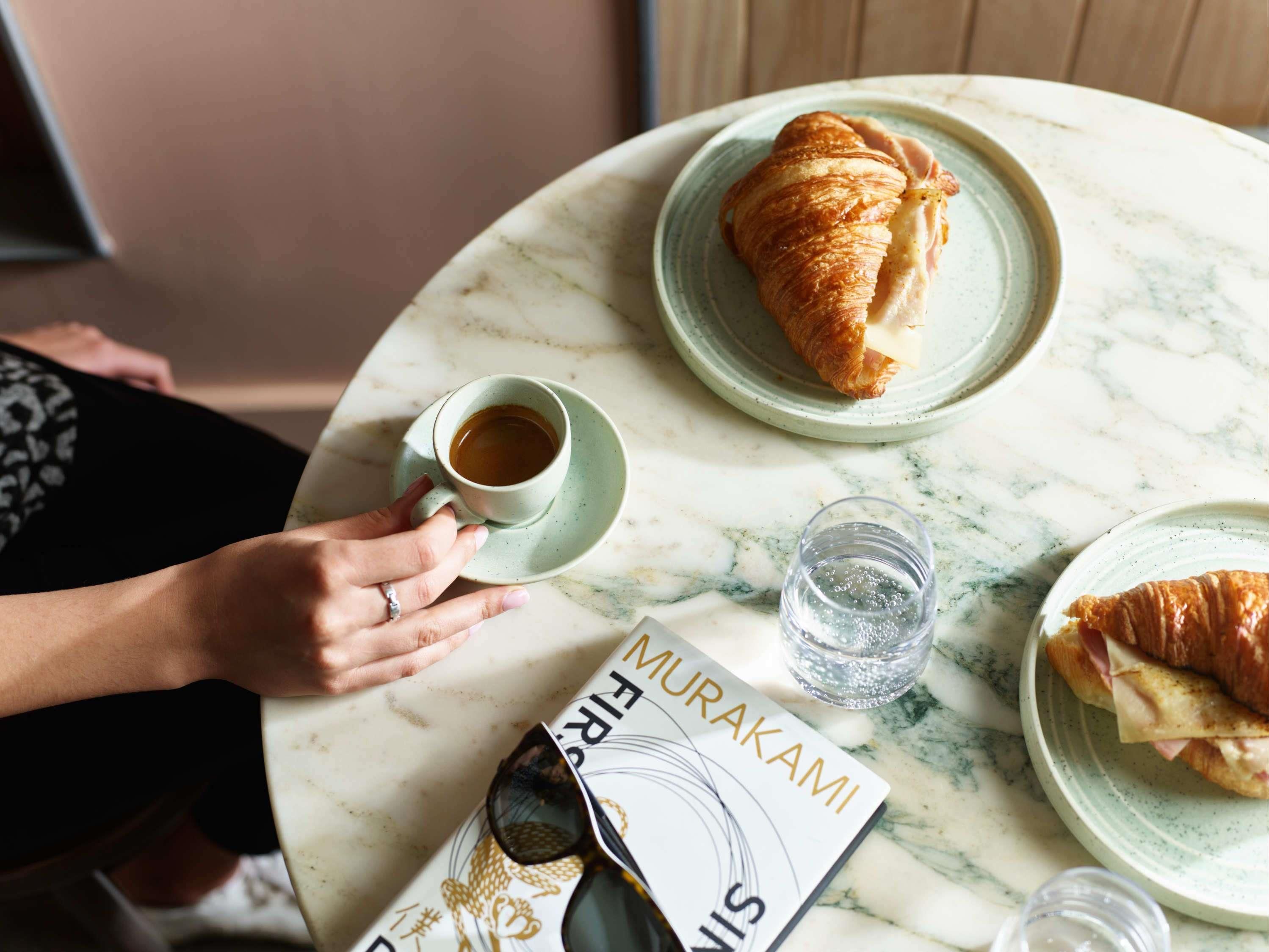 Una persona sosteniendo una taza de café, un croissant y un libro sobre una mesa de mármol.