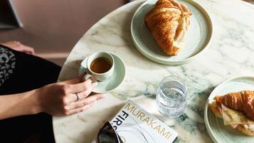 Una persona sosteniendo una taza de café, un croissant y un libro sobre una mesa de mármol.