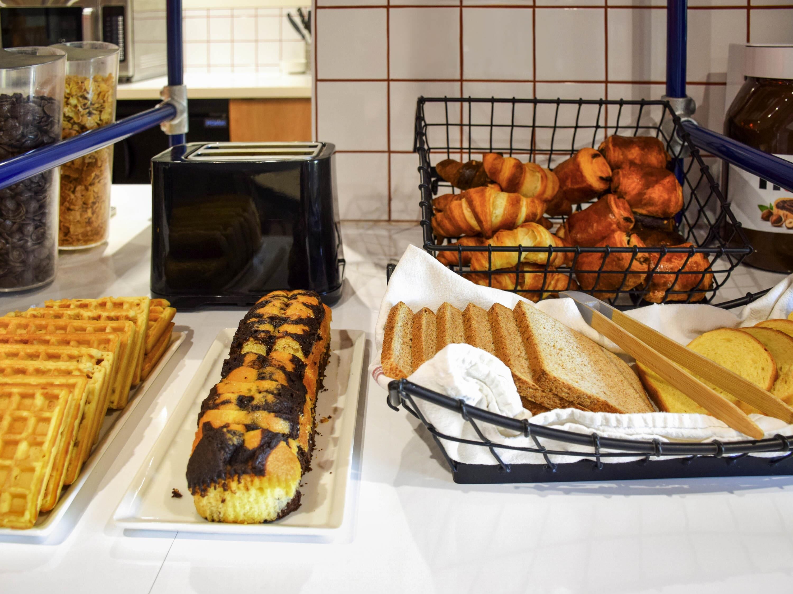 A buffet with various baked goods including waffles, a chocolate-topped loaf, croissants, and bread slices.