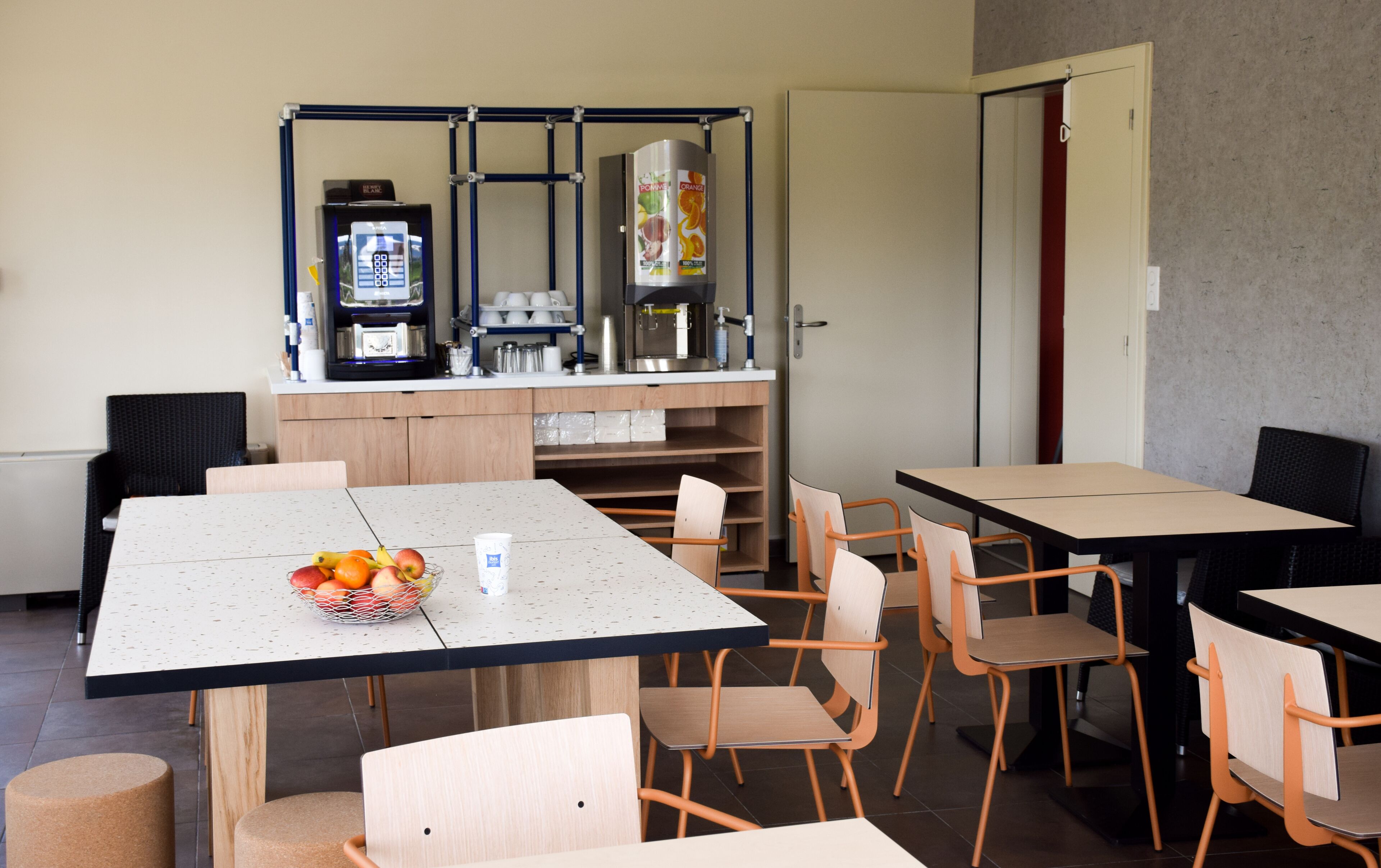 Un espace repas moderne avec des tables et des chaises, une machine à café et un panier de fruits posé sur la table.