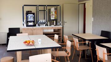 Un espace repas moderne avec des tables et des chaises, une machine à café et un panier de fruits posé sur la table.