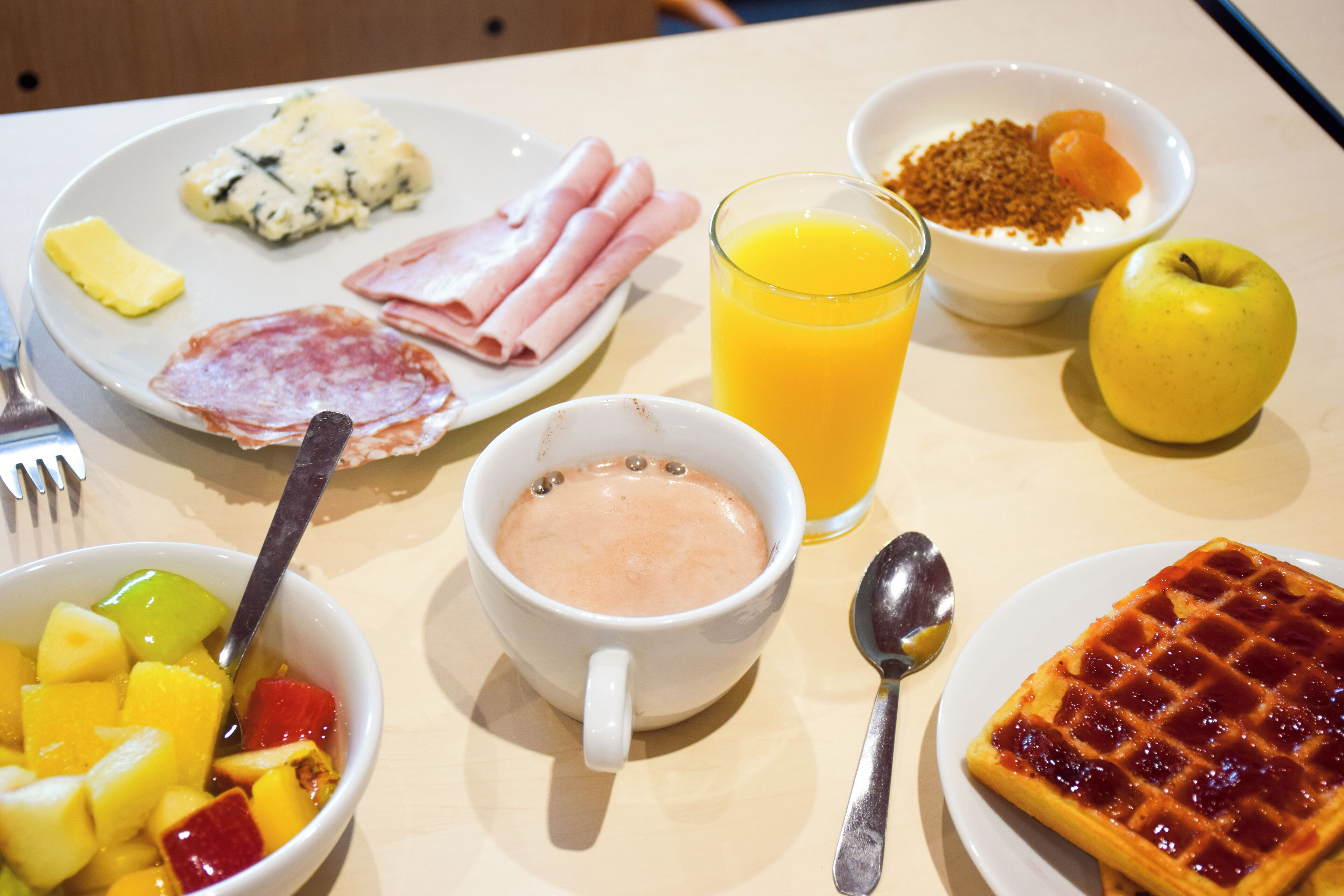 Une table de petit-déjeuner avec divers aliments, notamment une gaufre, des fruits, un verre de jus d’orange et un bol de yaourt.