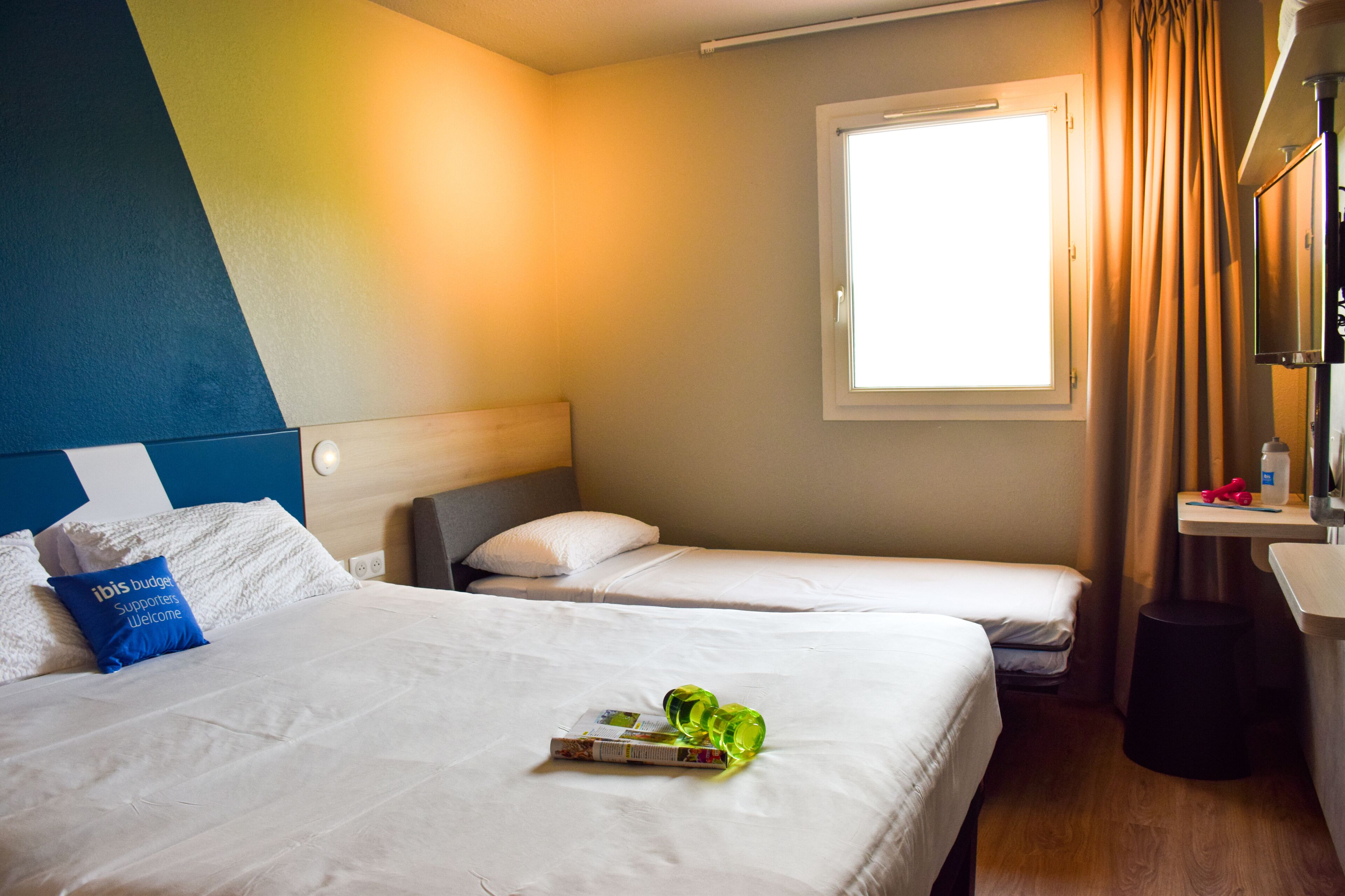 Camera d'albergo con due letti, una finestra con tende, un comodino e uno sgabello.