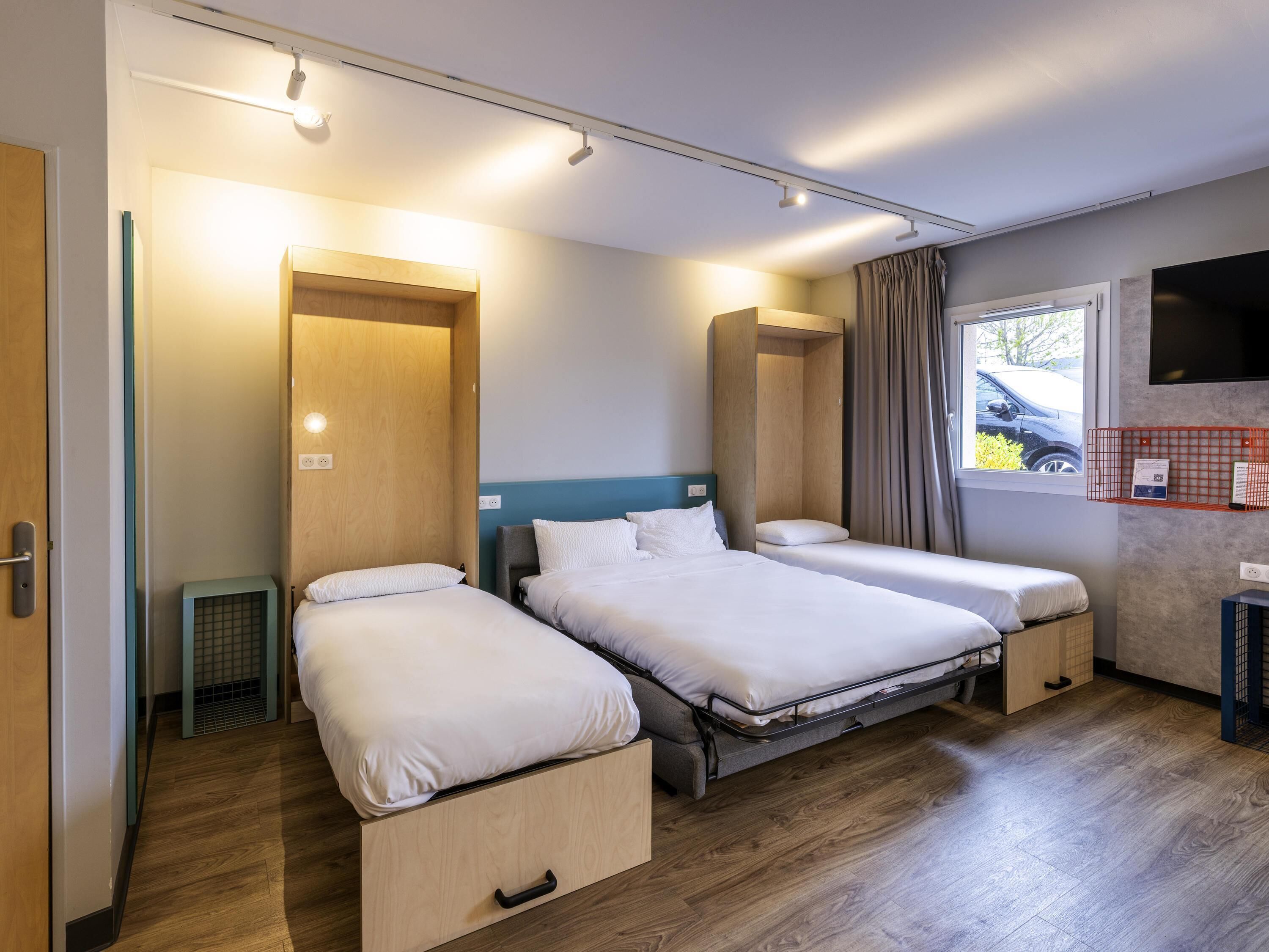 A hotel room with two beds, a wooden wardrobe, a small heater, a TV on the wall, and a window with curtains.