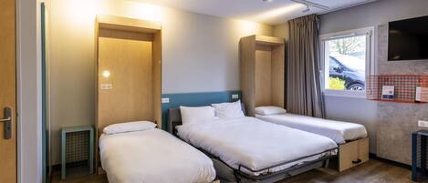 A hotel room with two beds, a wooden wardrobe, a small heater, a TV on the wall, and a window with curtains.