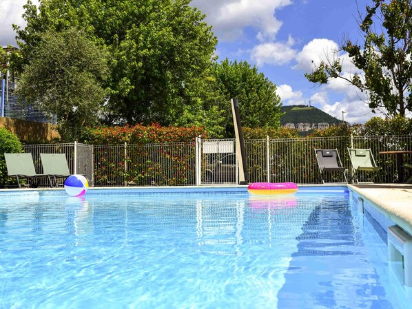 Una piscina con agua azul cristalina, rodeada por una valla metálica y árboles al fondo.