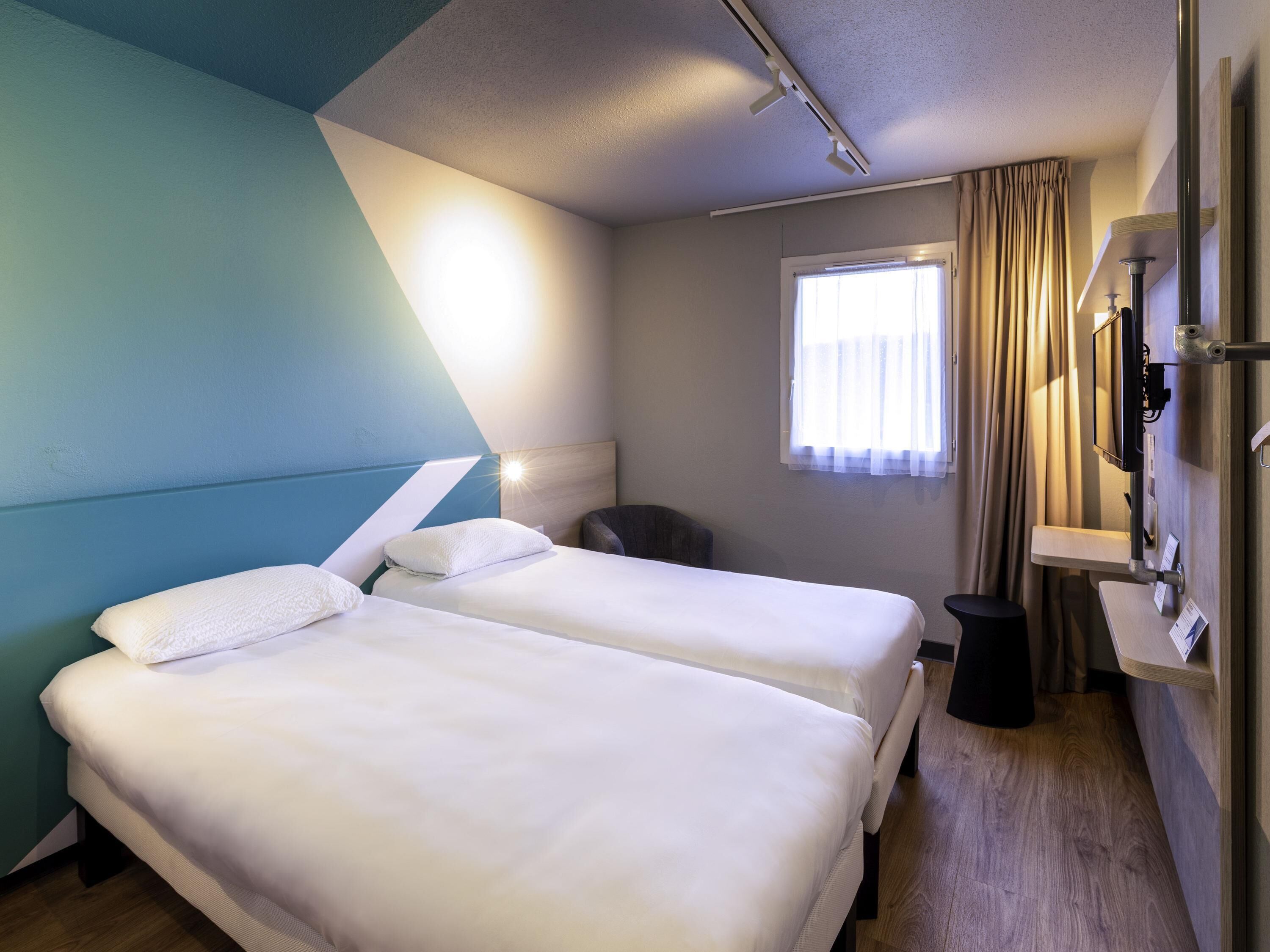 A hotel room with two beds, a window with curtains, a bedside table, and a mirror.