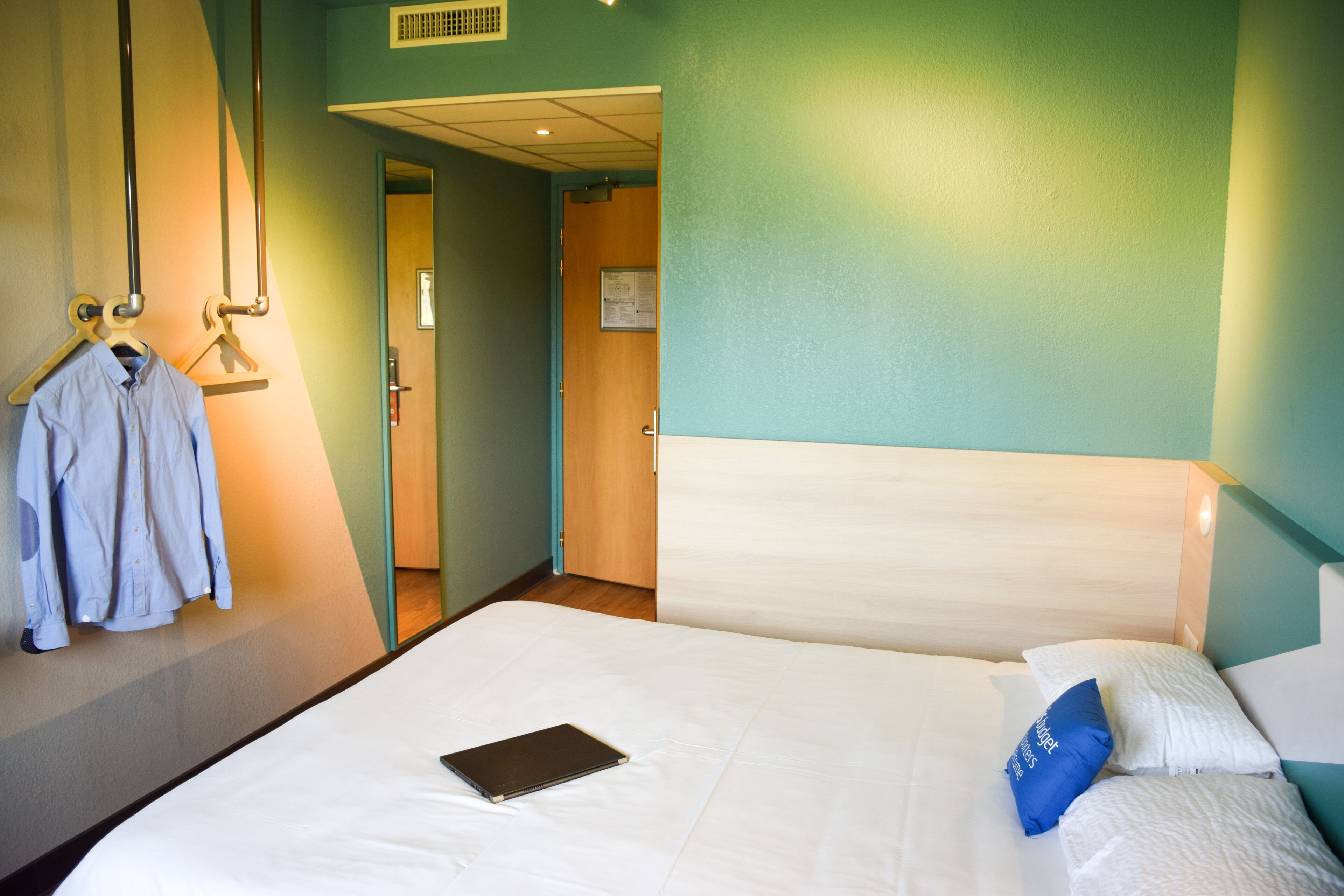Ein Hotelzimmer mit einem Bett, einem blauen Kissen, einem weißen Kissen, einem blauen Hemd an einem Kleiderbügel und einer Tür zu einem anderen Raum.