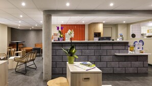 A reception area with a brick counter, a reception desk, and a seating area with chairs and a table. - Ibis Budget Millau Viaduc  (Millau)