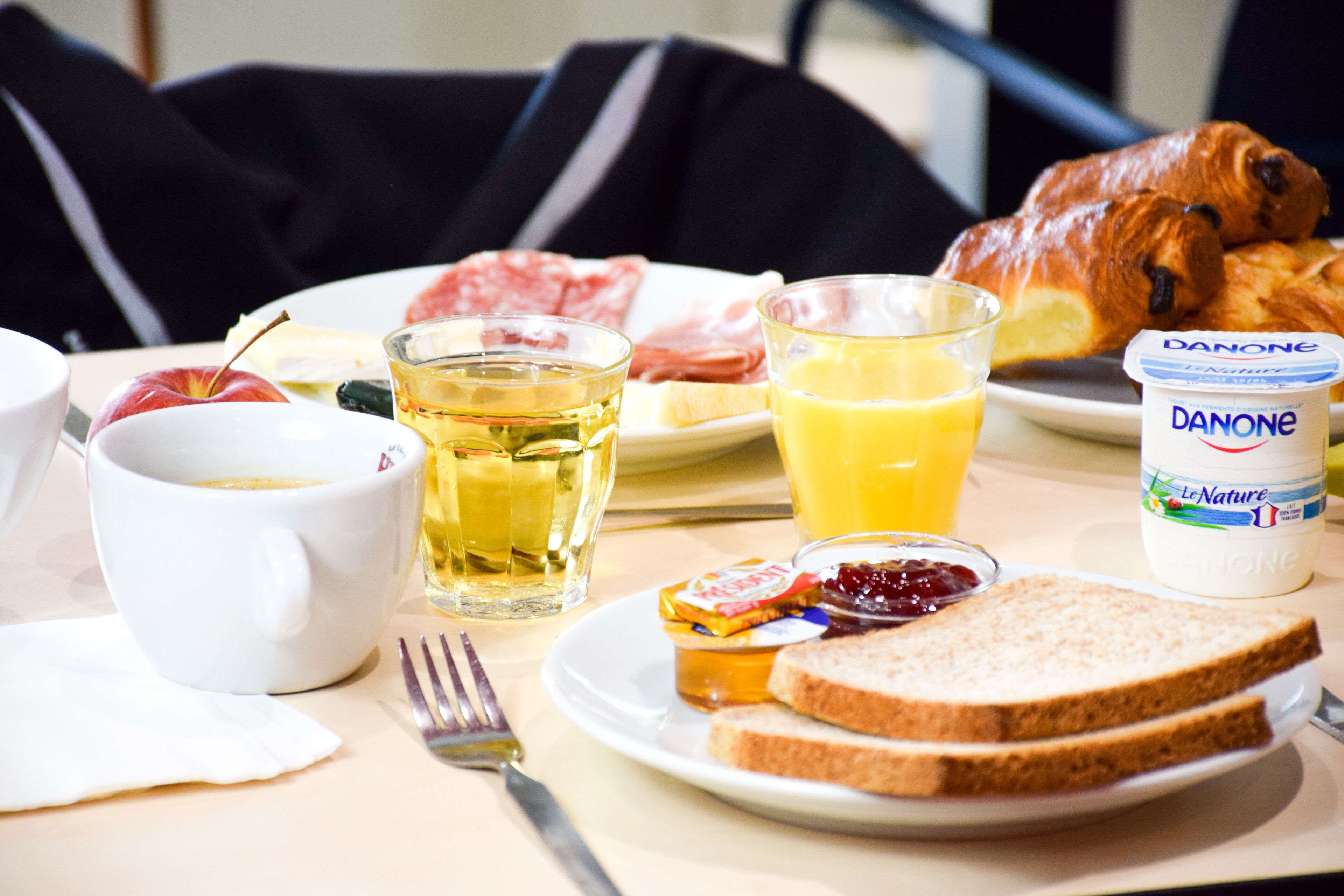 Un desayuno con croissants, un plato de jamón, un vaso de zumo de naranja, una taza de té y un envase de Danone.