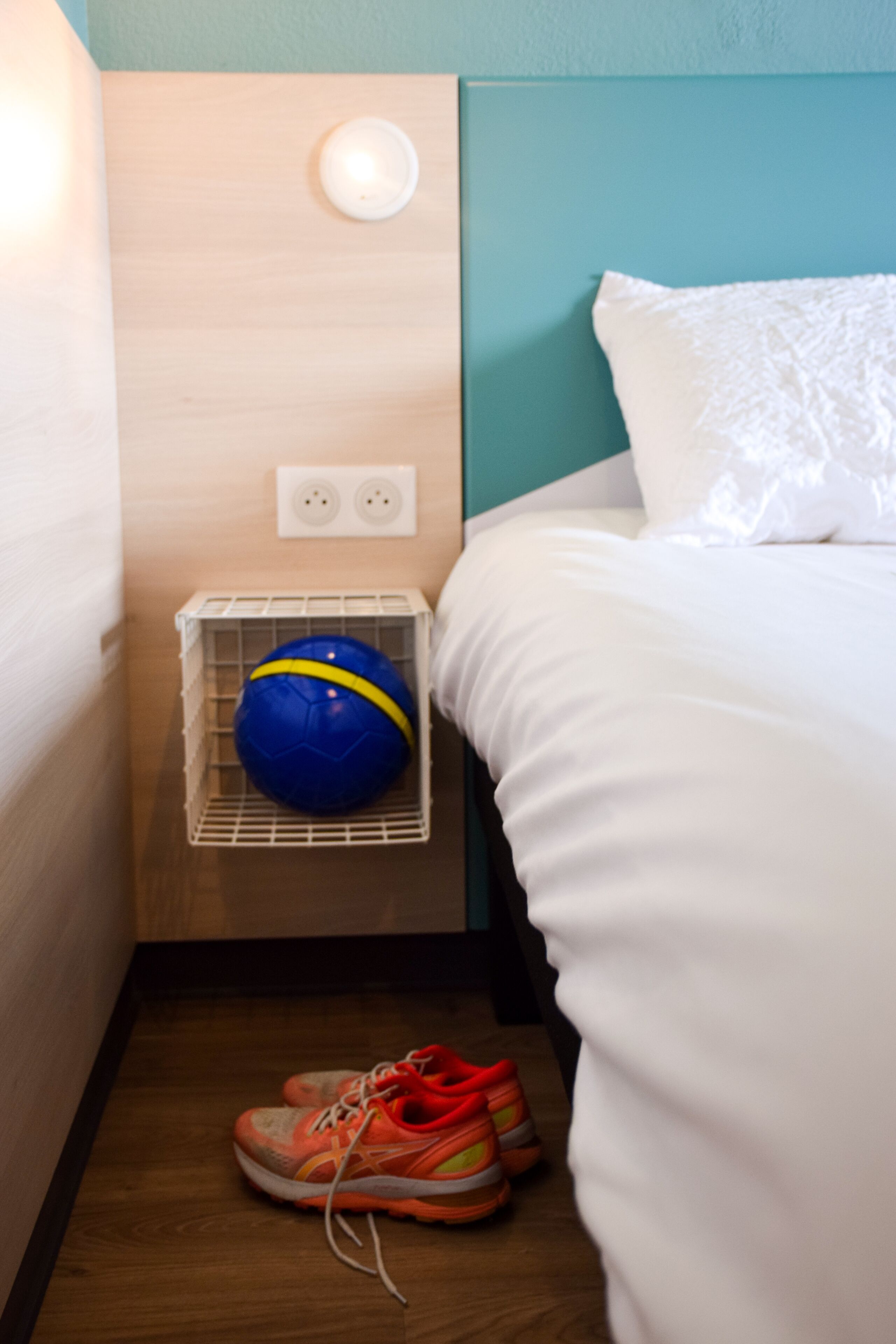 Una cama bien hecha con sábanas blancas, una pelota deportiva azul y amarilla en una cesta, y un par de zapatillas anaranjadas y blancas sobre el suelo.