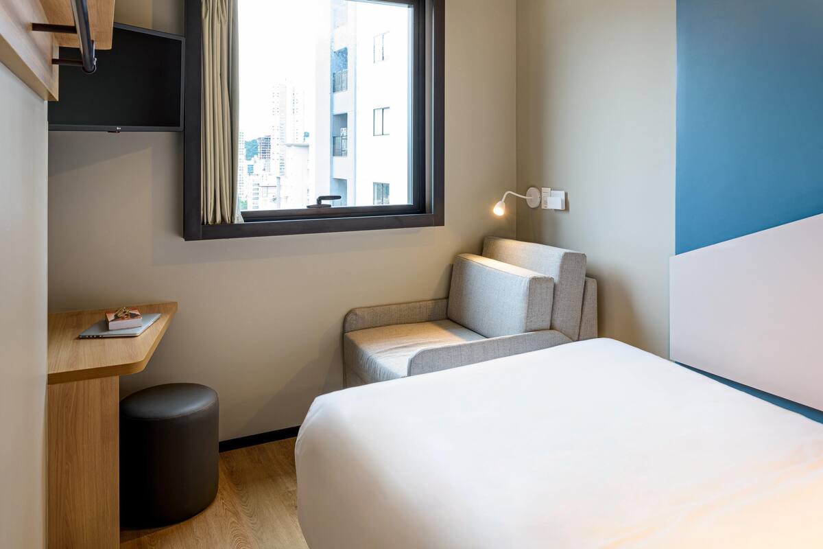 Een hotelkamer met een bed, een bureau, een stoel, een klein krukje, een raam met uitzicht op gebouwen en een wandlamp.