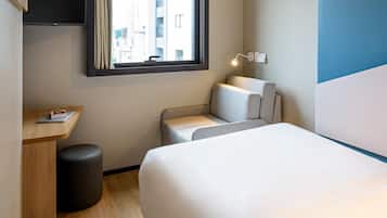 Quarto de hotel com cama, escrivaninha, cadeira, um banquinho pequeno, janela com vista para edifícios e luminária fixada na parede.