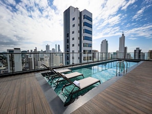 Une piscine sur le toit, dotée de chaises longues, d’une terrasse en bois et offrant une vue panoramique sur la ville.