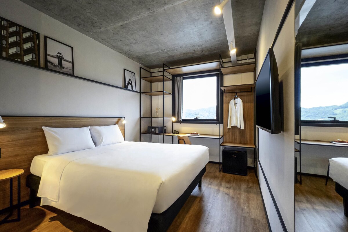 A modern hotel room with a large bed, bedside tables, a desk, a TV, and a window with a view.
