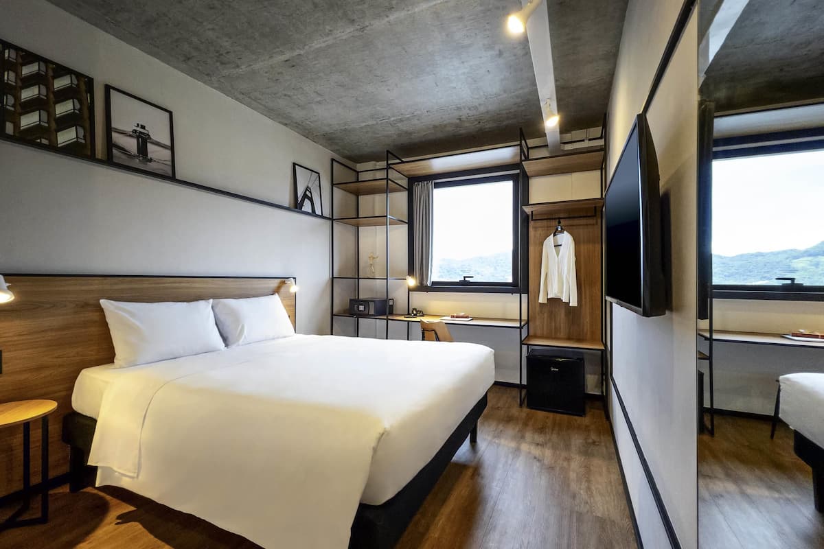 A modern hotel room with a large bed, bedside tables, a desk, a TV, and a window with a view.