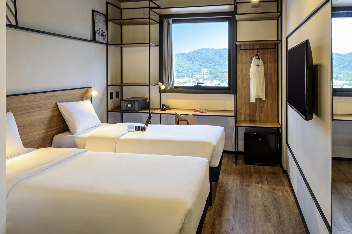 A hotel room with two beds, a desk, a chair, a TV, and a window with a view of the mountains.