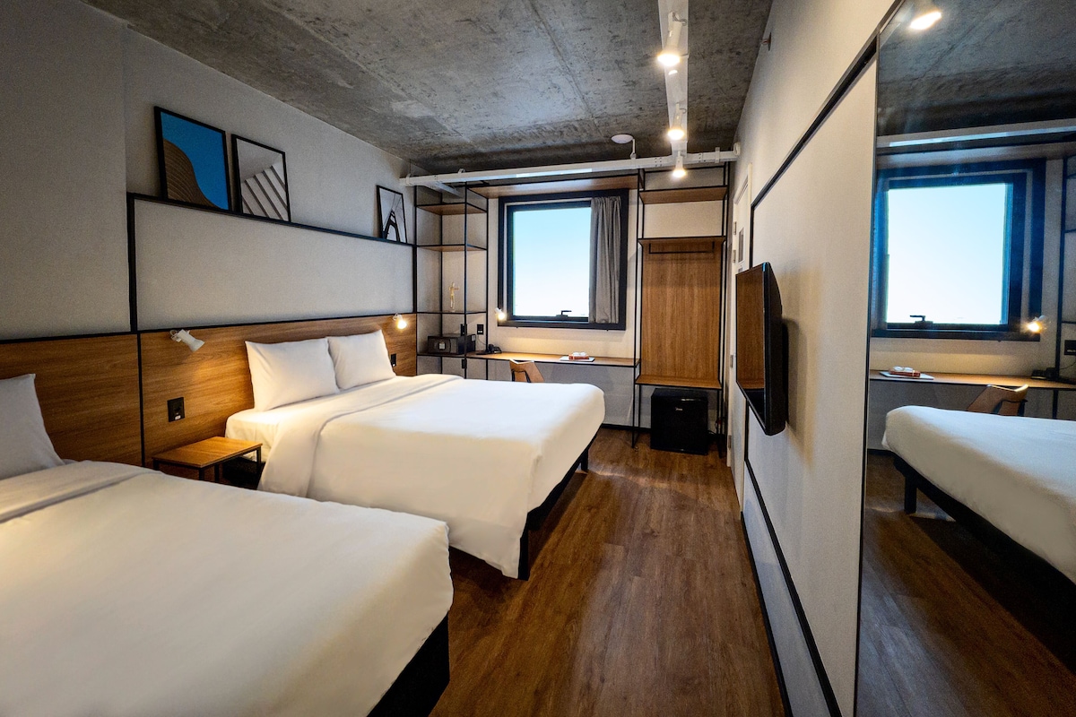 Habitación de hotel con dos camas, piso de madera, un ventanal grande y un televisor.
