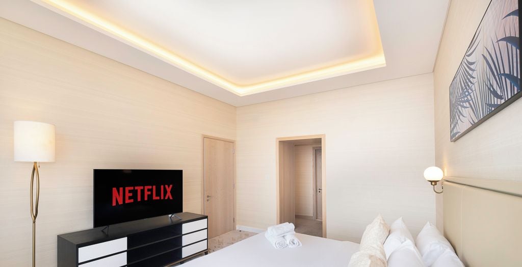 A modern hotel room with a large bed, a flat-screen TV displaying 'NETFLIX', a bedside lamp, and a wall-mounted artwork.