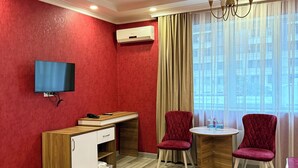 A room with a red wall, a flat-screen TV, a wooden desk, two red chairs, a chandelier, and a large window with curtains.