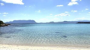 Ein Strand mit klarem Wasser, sandigem Ufer und Bergen im Hintergrund.