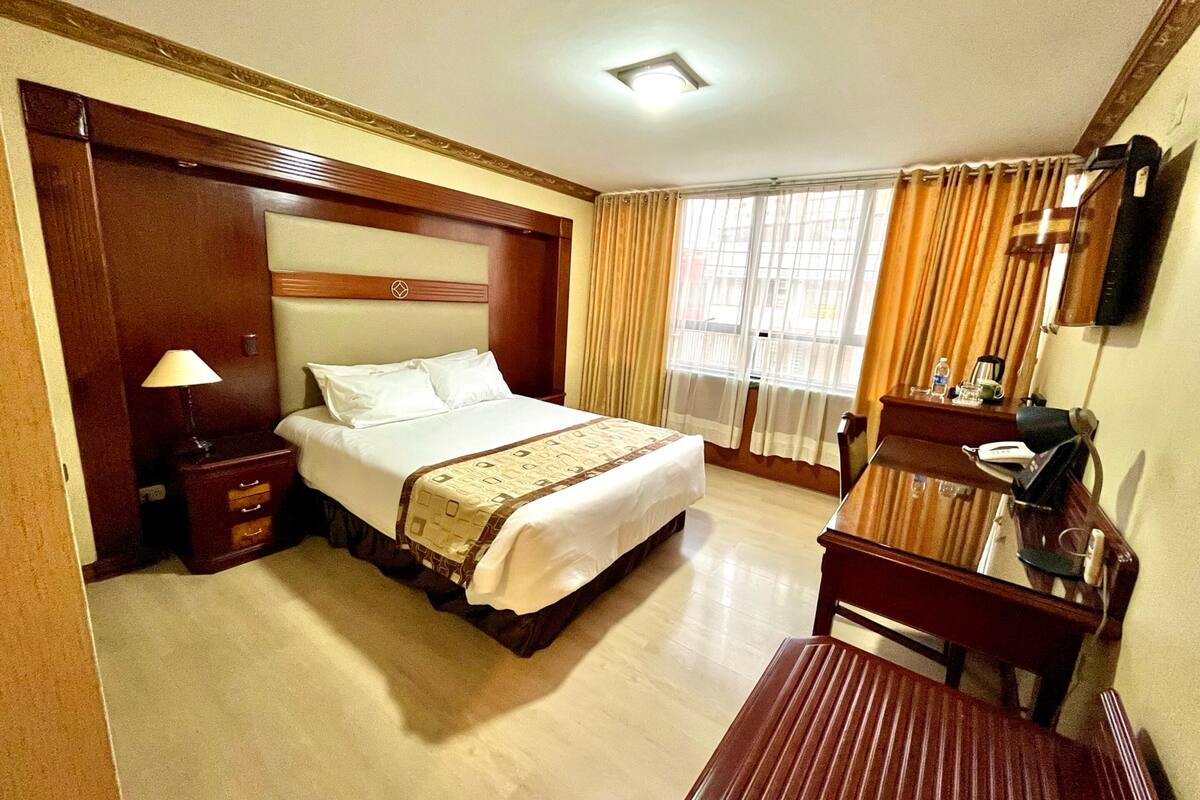 A hotel room with a bed, bedside table, lamp, desk, chair, and a TV mounted on the wall.