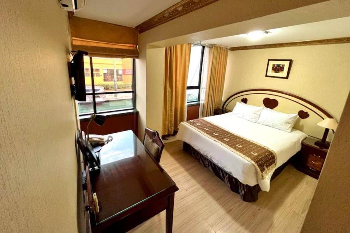 A hotel room with a bed, a desk, a chair, a window with curtains, and a picture on the wall.