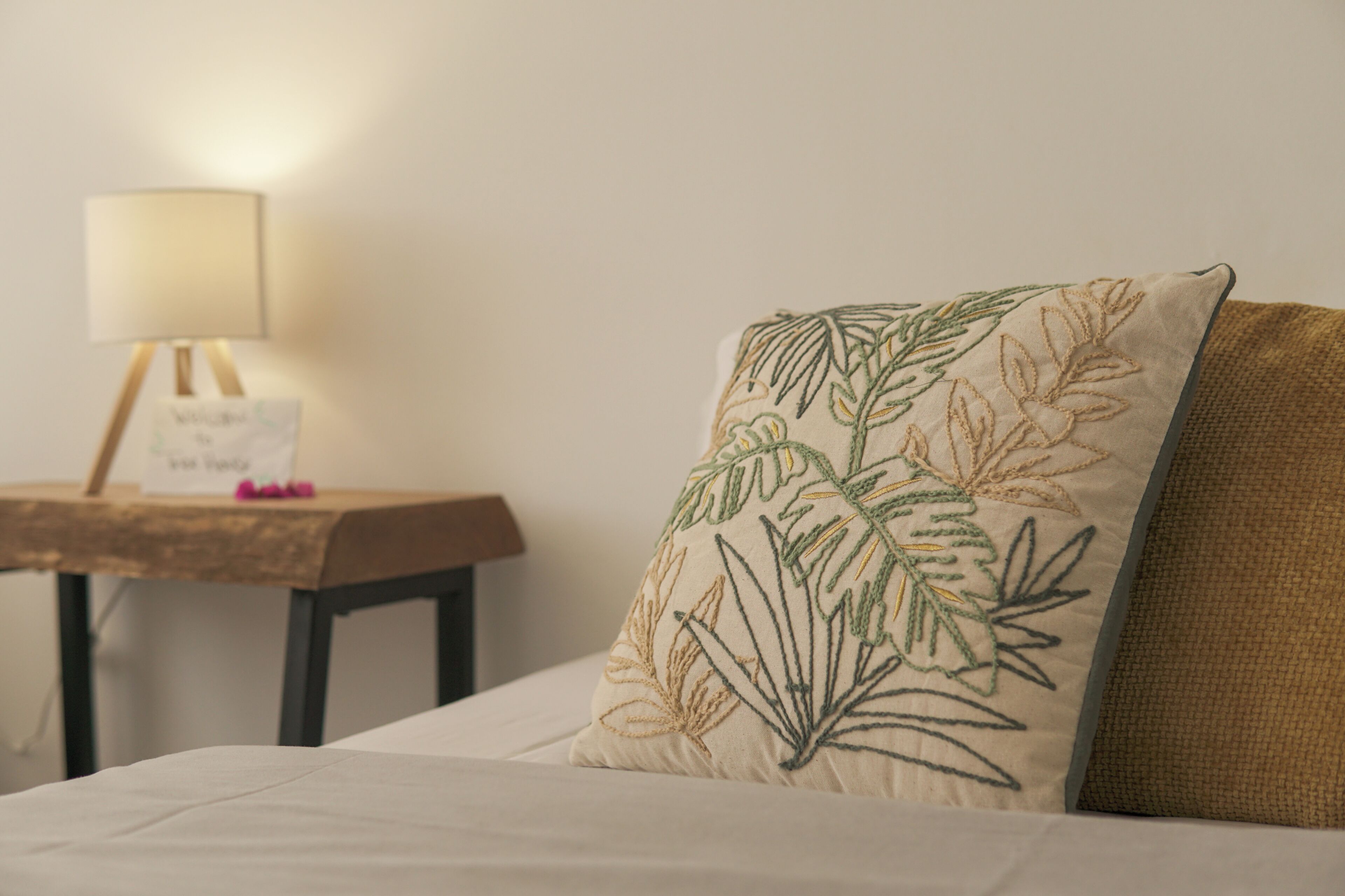 A bed with a pillow featuring a leaf pattern, a bedside table with a lamp, and a sign that reads 'Welcome to [Hotel Name].'