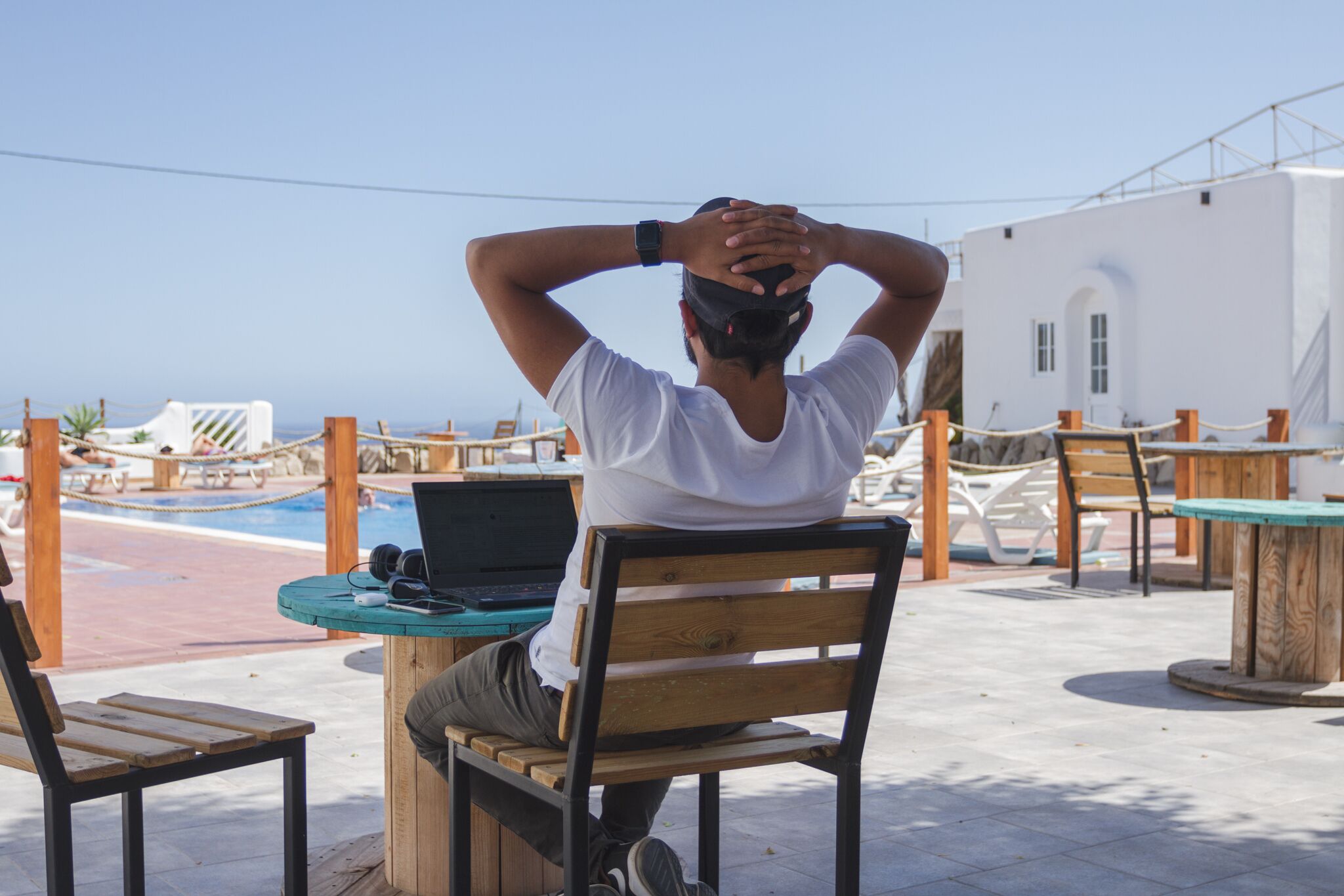A person sitting at a wooden table with a laptop, overlooking a pool area with lounge chairs and a white building in the background.