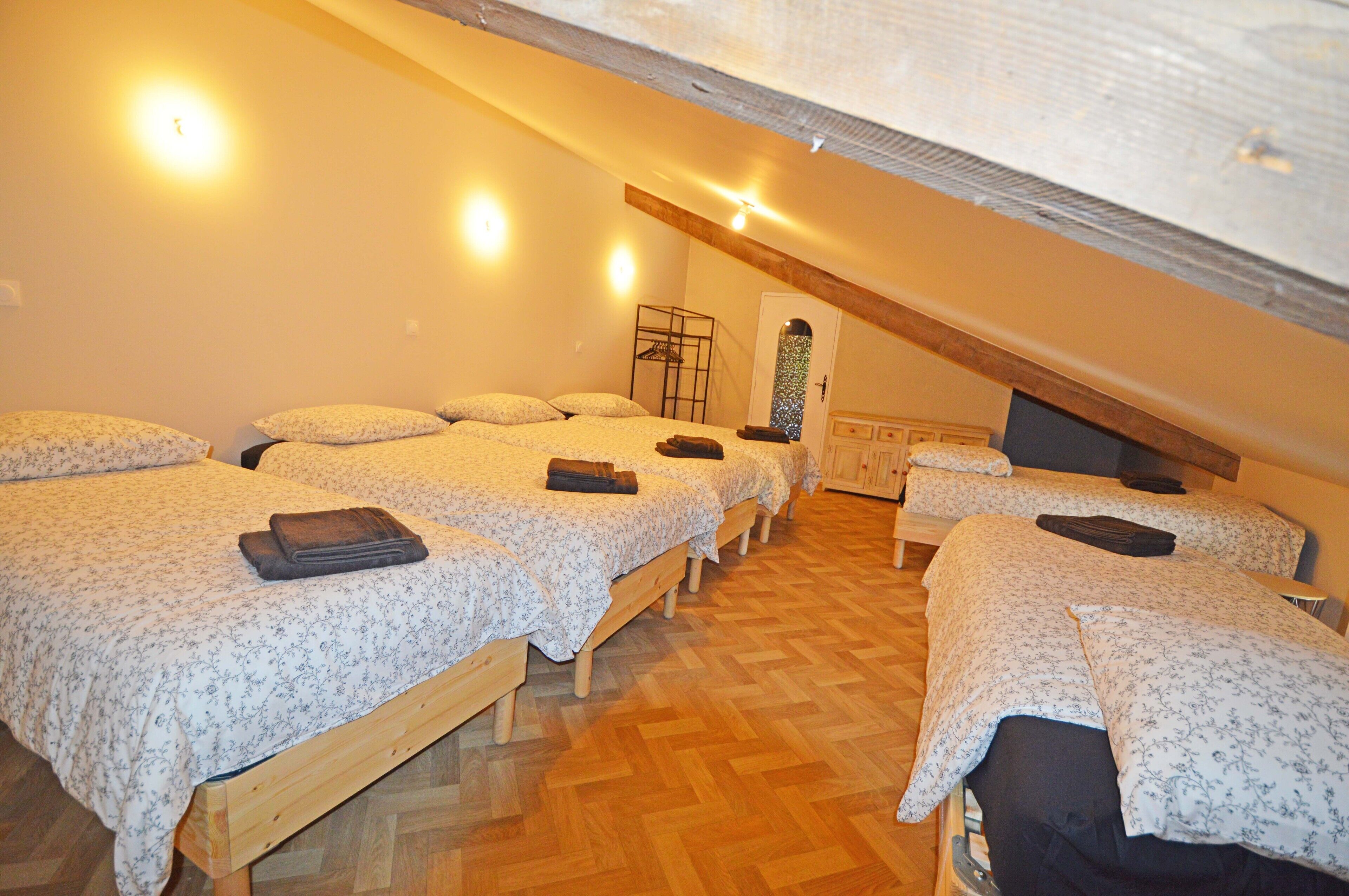 A room with three wooden beds, each with a floral bedspread and pillows. There is a wooden dresser and a small table with a chair. The room has wooden flooring and a sloped ceiling with exposed beams.