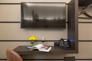 A hotel room with a flat-screen TV mounted on the wall, a desk with a book, a vase of yellow flowers, a wine glass, and a telephone. - Palace Hotel Due Ponti (Siena)