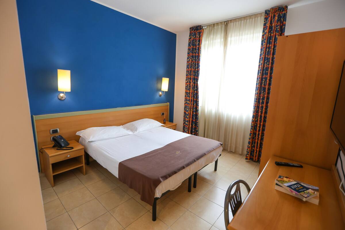 A hotel room with a bed, bedside table, lamp, and a desk with a book.