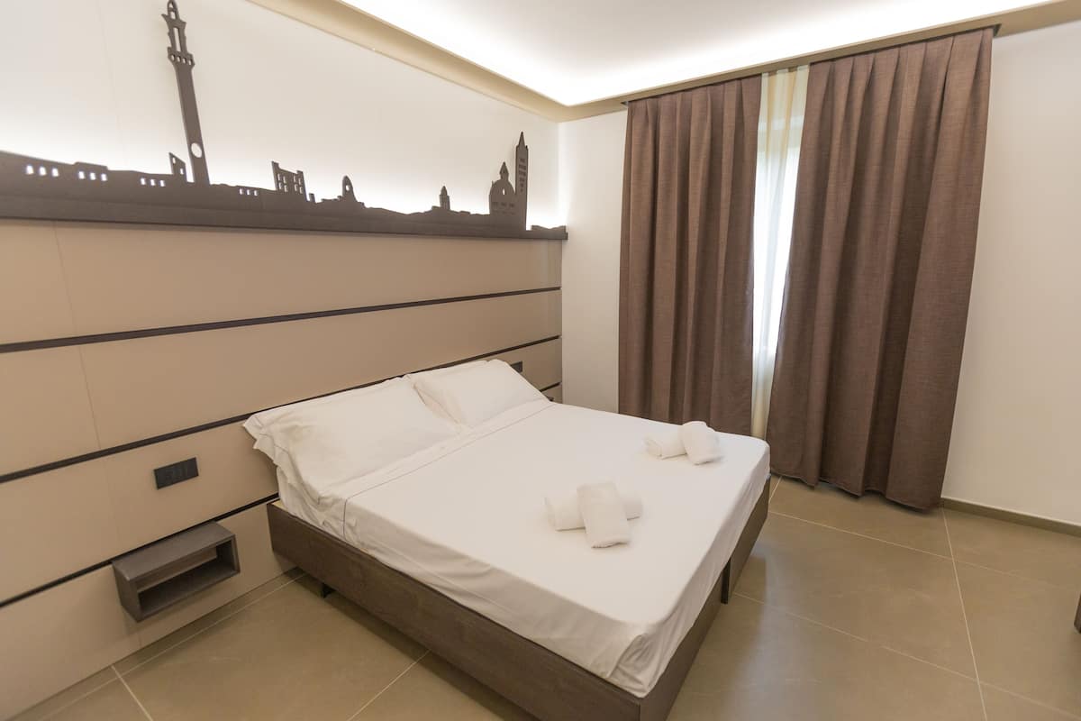 A neatly made bed with white linens and towels, a cityscape silhouette on the wall, a window with curtains, and a built-in shelf.