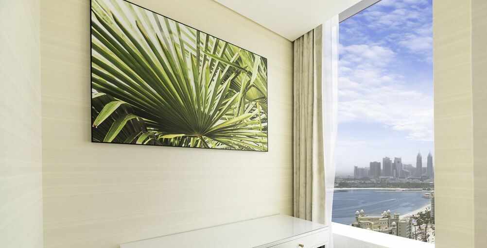 A modern room with a large window, a city skyline view, and a wall-mounted artwork of palm leaves.