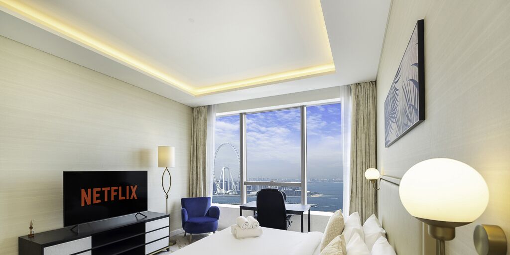 A hotel room with a large bed, a desk, a chair, a TV displaying 'NETFLIX', a lamp, and a view of a bridge and water.