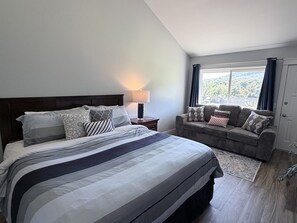 A hotel room with a large bed, a sofa, a nightstand with a lamp, and a view of the outdoors through a window. - Fairway Villa 604 at Rumbling Bald (Lake Lure)