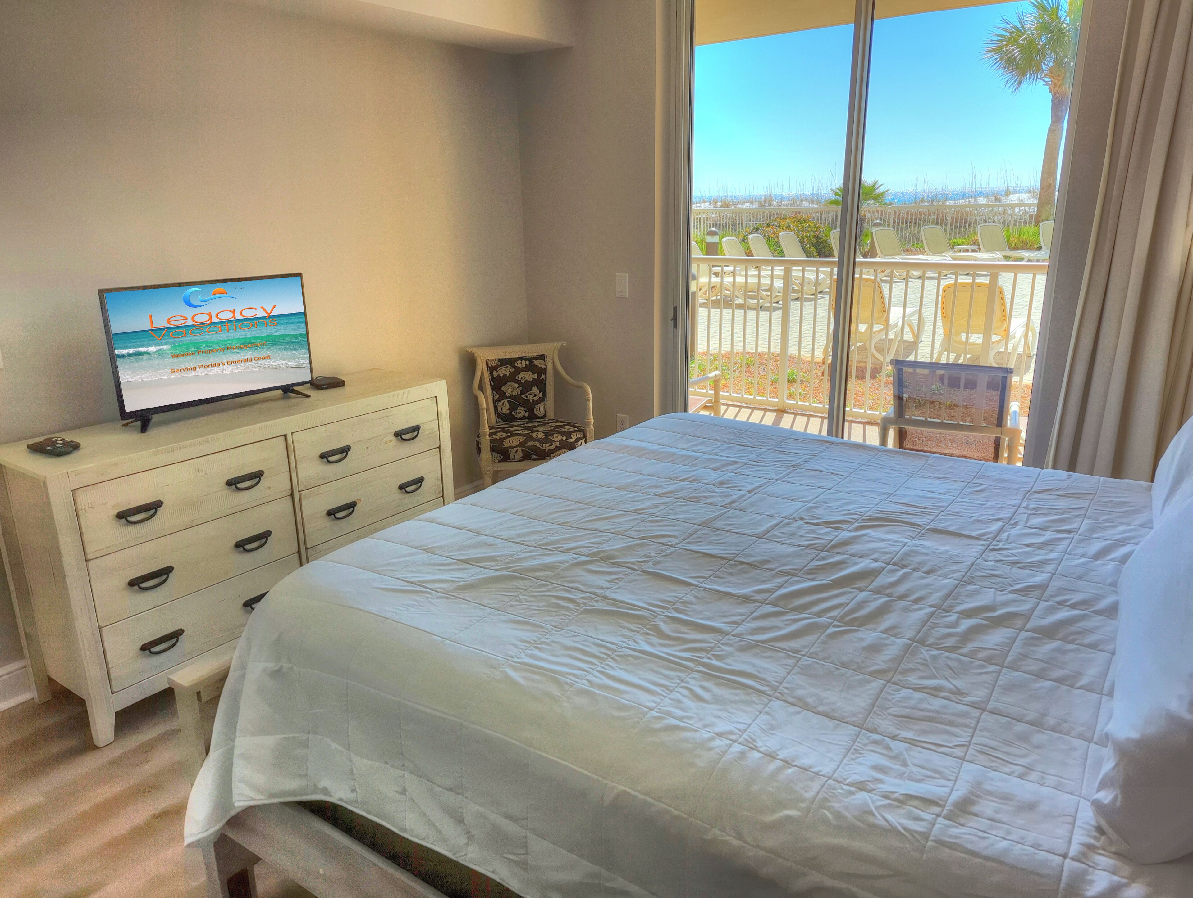 A hotel room with a bed, a dresser, a television displaying 'Legacy Vacations', and a balcony with chairs and a view of the beach.