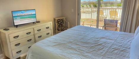 A hotel room with a bed, a dresser, a television displaying 'Legacy Vacations', and a balcony with chairs and a view of the beach.