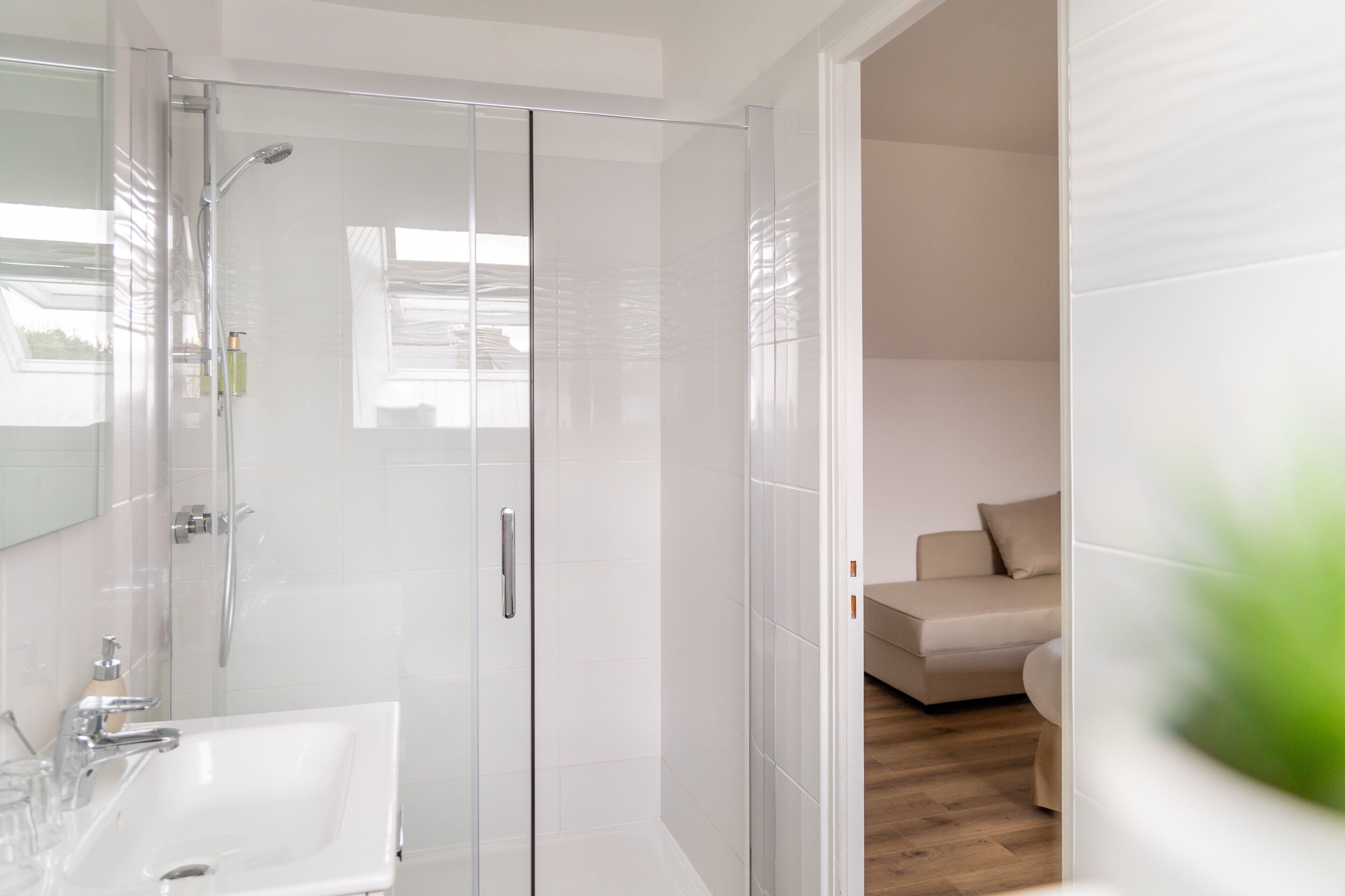A modern bathroom with a glass-enclosed shower, a white sink, and a beige sofa in the adjacent living area.