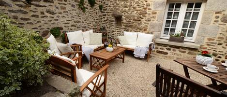 A stone-walled courtyard with wooden furniture, including sofas and a table, and a window with a flower box.